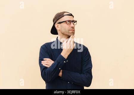 Thoughtful young man in glasses touches his chin while standing against beige wall outdoors Stock Photo