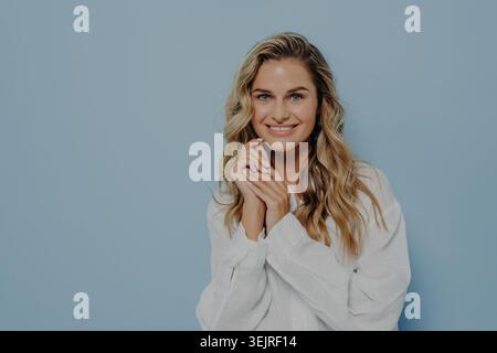 Smiling young blonde woman poses in white shirt against blue studio wall Stock Photo
