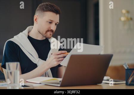 Young man reviews documents while working on laptop at home office desk Stock Photo