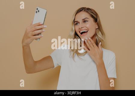 Young woman taking selfie with smartphone against beige studio wall Stock Photo