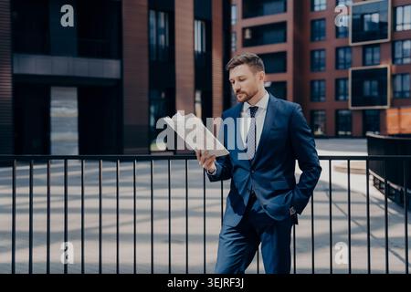 Businessman in blue suit reading newspaper outside modern office buildings Stock Photo