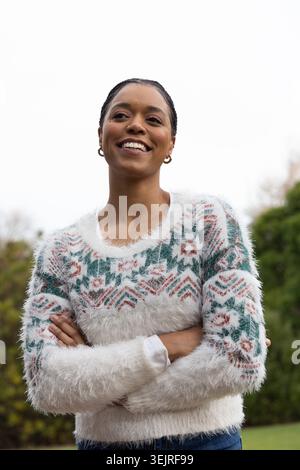Smiling, standing with arms crossed in garden, African American woman wearing fuzzy sweater. Portrait, outdoor, knitwear, geometric, pattern, lawn, ov Stock Photo