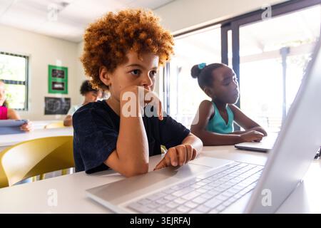 Diverse elementary students working on laptops and tablets at classroom desks with yellow chairs. Pupils, learning, education, d Stock Photo