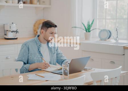 Man working on laptop and writing notes at kitchen table in bright home interior Stock Photo
