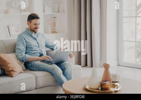 Young man works on laptop while sitting on sofa in bright living room Stock Photo