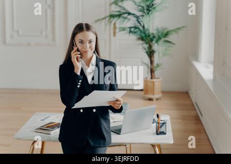 Businesswoman talks on smartphone while holding documents in bright modern office Stock Photo