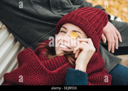 Young woman in red knit hat lying on bench and holding autumn leaf Stock Photo