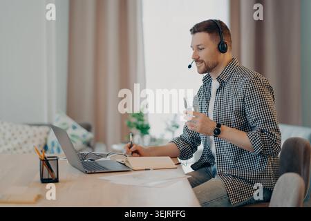 Man wearing headset taking notes while working on laptop at home office desk Stock Photo