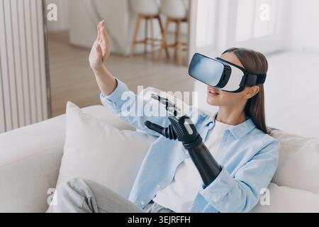 Woman with bionic hand interacts in virtual reality while relaxing in modern living room Stock Photo