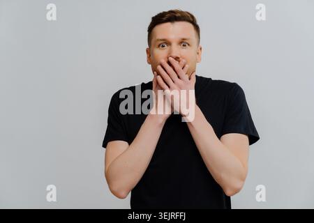 Shocked young man covering mouth with hands against gray studio backdrop Stock Photo