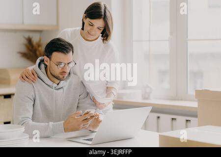Young couple reviewing bills on laptop while moving into new apartment Stock Photo