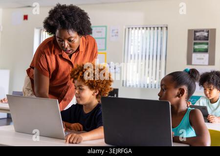 African American teacher at school leaning over child using laptop, peers using laptops and tablet. Educator, learners, devices, Stock Photo