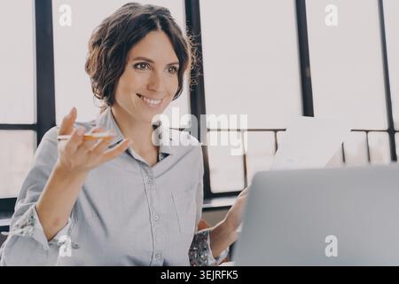 Woman presents business documents while sitting at laptop near office windows Stock Photo