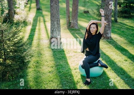 Woman stretches on exercise ball in green park during outdoor fitness workout Stock Photo