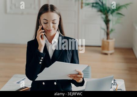 Professional woman in black suit reads paperwork during business phone call Stock Photo