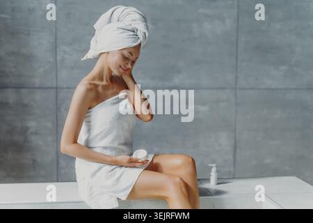 Young woman applies face cream while wrapped in towel in modern spa bathroom Stock Photo