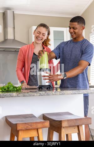 Diverse couple preparing green smoothie at kitchen island with blender, wearing coral jacket. Drink, herbs, parsley, cilantro, h Stock Photo