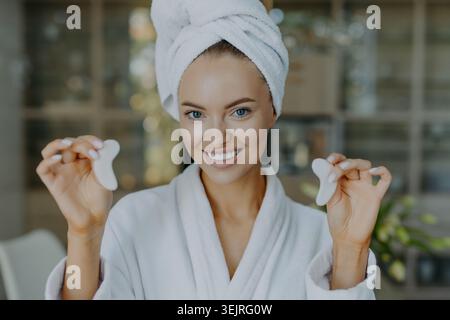 Smiling woman demonstrates white facial massage tools while wearing towel turban Stock Photo