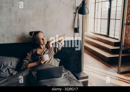 Young woman drinks coffee while using laptop in bed in loft apartment Stock Photo