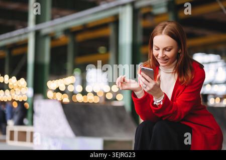 Smiling red-haired woman uses smartphone in modern urban public space with lights Stock Photo