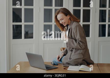 Smiling female freelancer planning work with laptop and pen in bright room Stock Photo