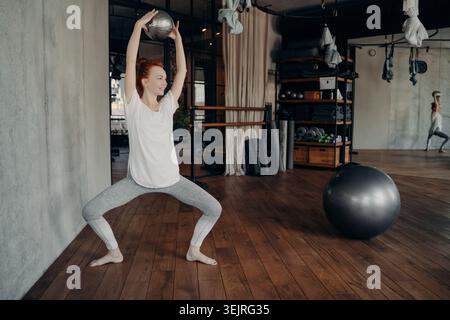 Redhead woman in wide squat position lifting medicine ball overhead and smiling in modern pilates studio with Swiss ball in back Stock Photo