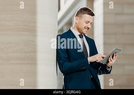 Businessman uses digital tablet while standing outside modern office building Stock Photo