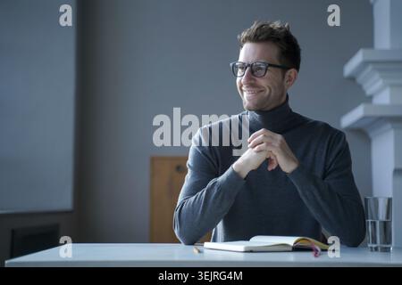 Smiling man in gray turtleneck sitting with notebook and water glass at home table Stock Photo