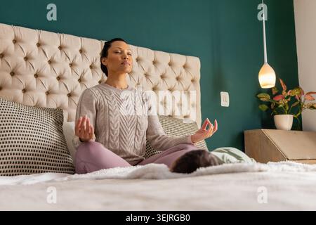 Mid adult woman meditating on bed in sweater and leggings with infant near headboard, lamp. Mindfulness, serenity, relaxation, m Stock Photo