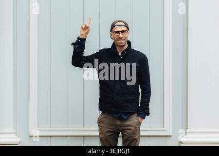 Middle aged man wearing glasses pointing upward near pale exterior wall Stock Photo