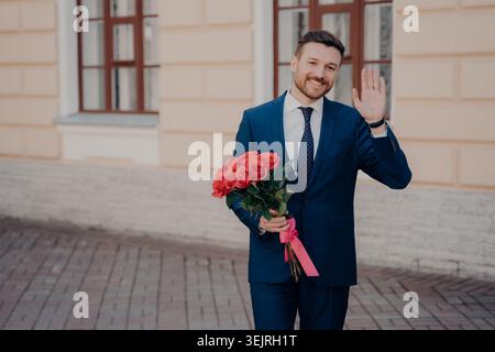 Young man in blue suit holding red roses and waving outdoors on city street Stock Photo