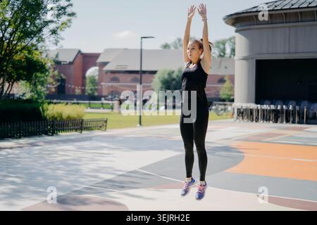 Young woman jumping with arms raised during outdoor fitness workout in urban park Stock Photo