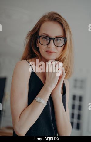 Red-haired young woman wearing black eyeglasses poses indoors with hands near face Stock Photo