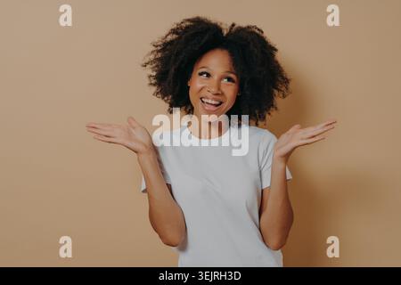 Young Black woman with curly afro hair smiling and presenting with open hands against beige studio wall Stock Photo