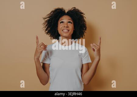Black woman in white t-shirt pointing upward while smiling against beige studio backdrop Stock Photo