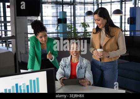 Diverse female team collaborating in workwear around table in office lounge with tablet and coffee. Workspace, collaboration, de Stock Photo