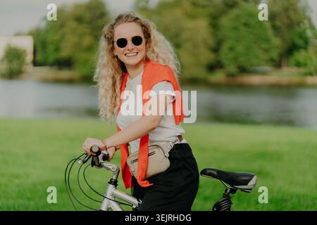 Young blonde woman smiles while standing with a bicycle near a lake in a green park Stock Photo