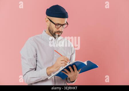 Middle aged man writes notes with pencil in blue notebook against pink studio wall Stock Photo