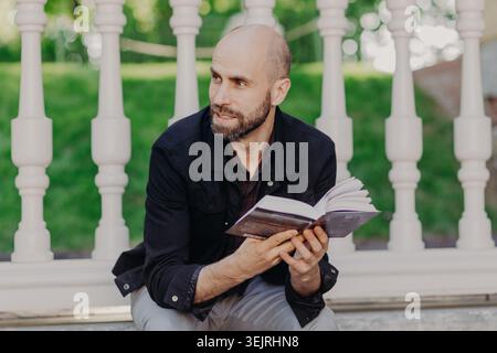 Bald bearded man reads a book while sitting on a porch in a green park Stock Photo