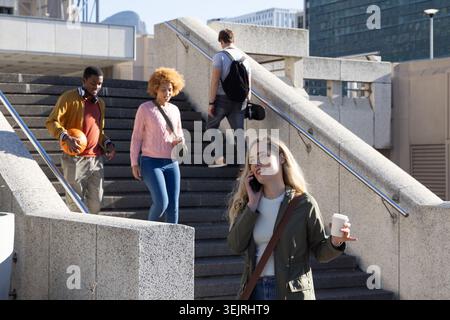 Diverse people descending concrete stairs, woman holding to-go coffee talking on phone, copy space. Urban, plaza, pedestrians, b Stock Photo
