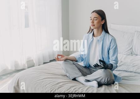 Young woman with prosthetic hand meditating on bed in bright bedroom Stock Photo