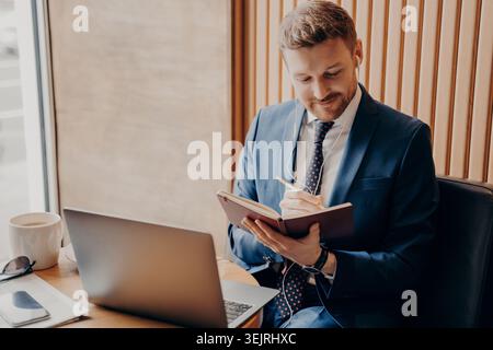 Businessman writes notes in planner beside laptop while working at cafe table Stock Photo