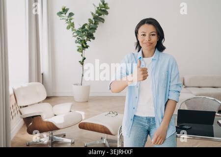 Young woman giving thumbs up beside laptop in modern home office Stock Photo
