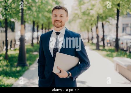Smiling businessman in blue suit holding laptop while standing on sunny city street Stock Photo
