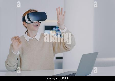 Young woman uses virtual reality headset and gestures beside laptop in modern office Stock Photo