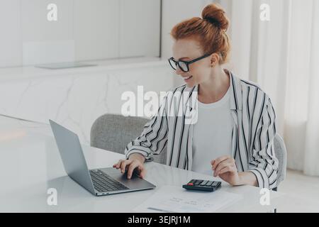Red-haired woman calculates finances on laptop with calculator and reports in modern home office Stock Photo