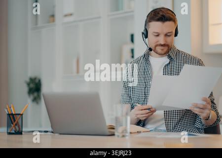 Male customer support agent wearing headset reviews documents beside laptop in home office Stock Photo