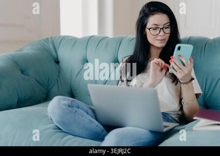 Dark-haired woman with glasses sitting cross-legged on teal sofa using smartphone while laptop is open on her lap at home Stock Photo