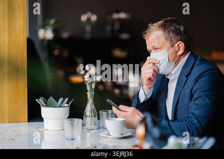 Businessman wearing medical mask uses smartphone while sitting alone at cafe table Stock Photo