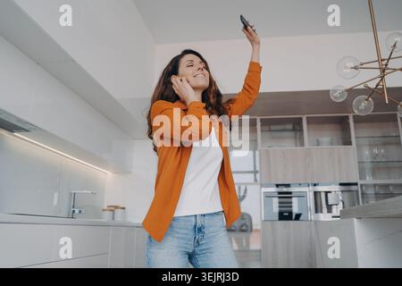 Young woman dances with smartphone while listening to music in modern kitchen at home Stock Photo